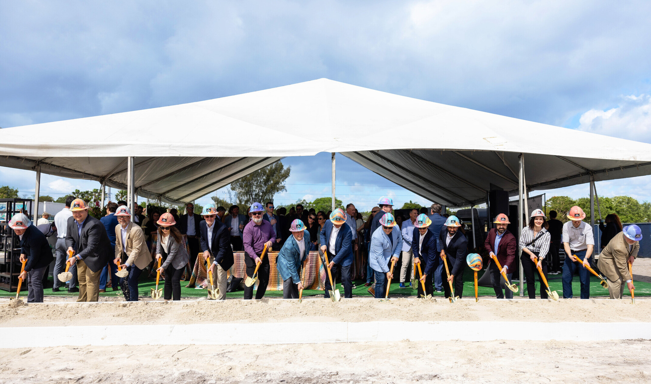 Multiple people in hard hats under a white tent holding shovels for a groundbreaking