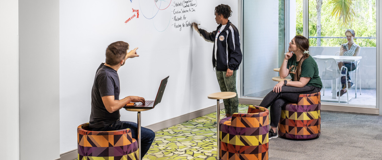 Student writing on a white board while interacting with others in a classroom with colorful seating