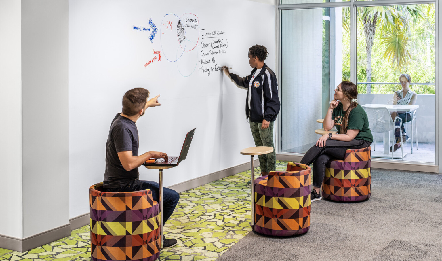 Student writing on a white board while interacting with others in a classroom with colorful seating