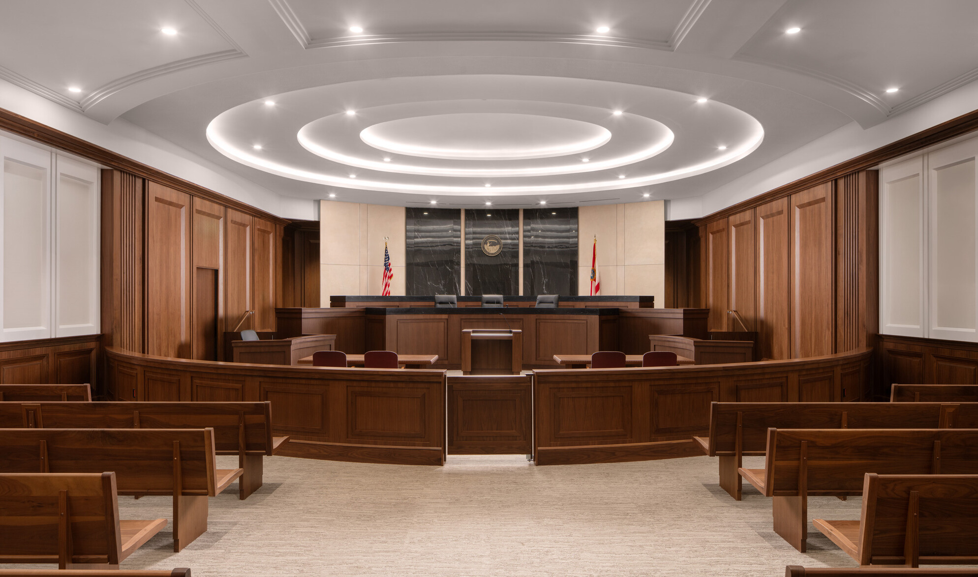 Interior courtroom image facing the judge’s bench; wood benches and barriers make for a official feel with a circular lighting element overtaking the ceiling