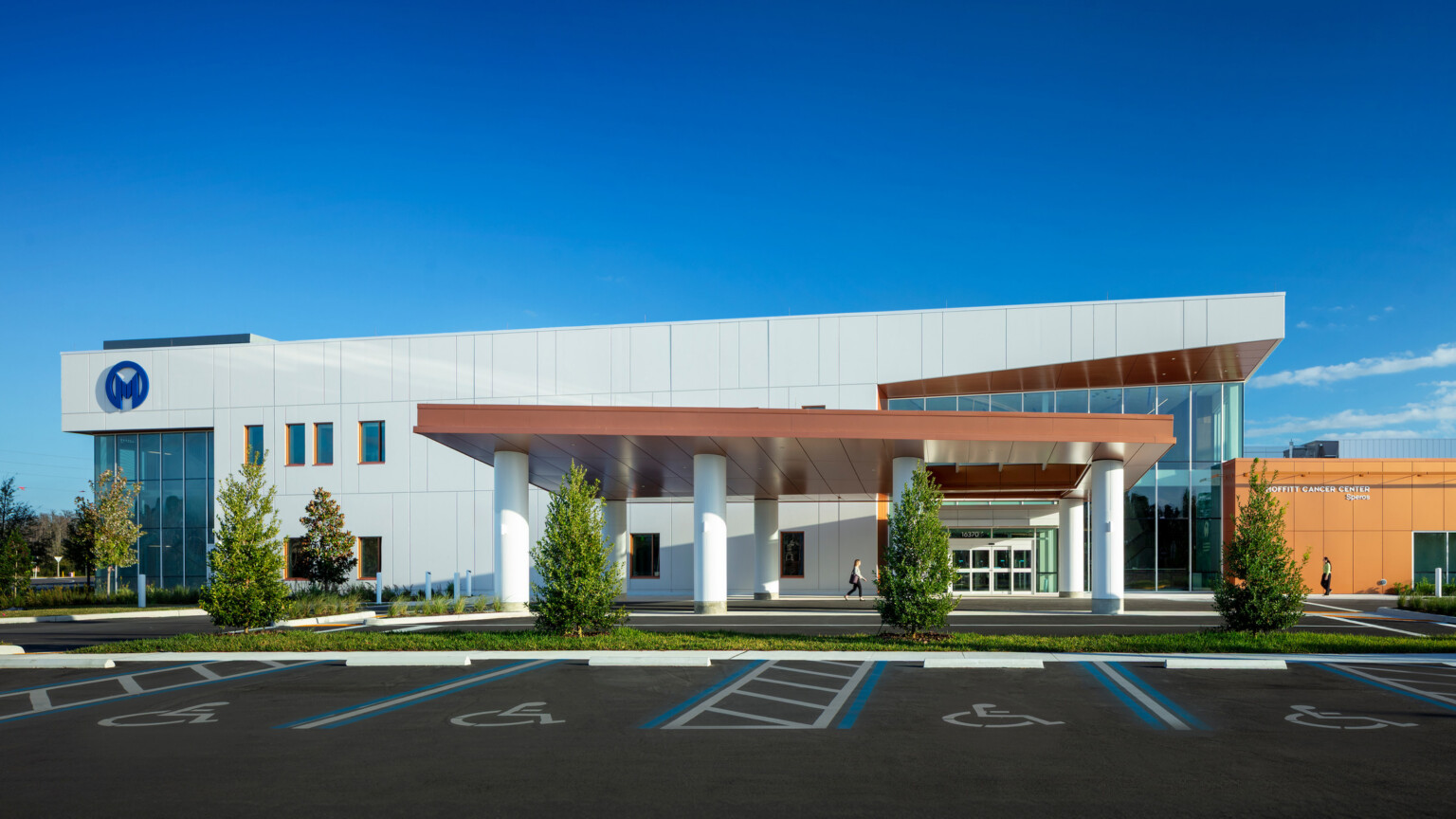 A modern building with a wide covered entrance, landscaped trees, and accessible parking spaces in the foreground under a bright blue sky