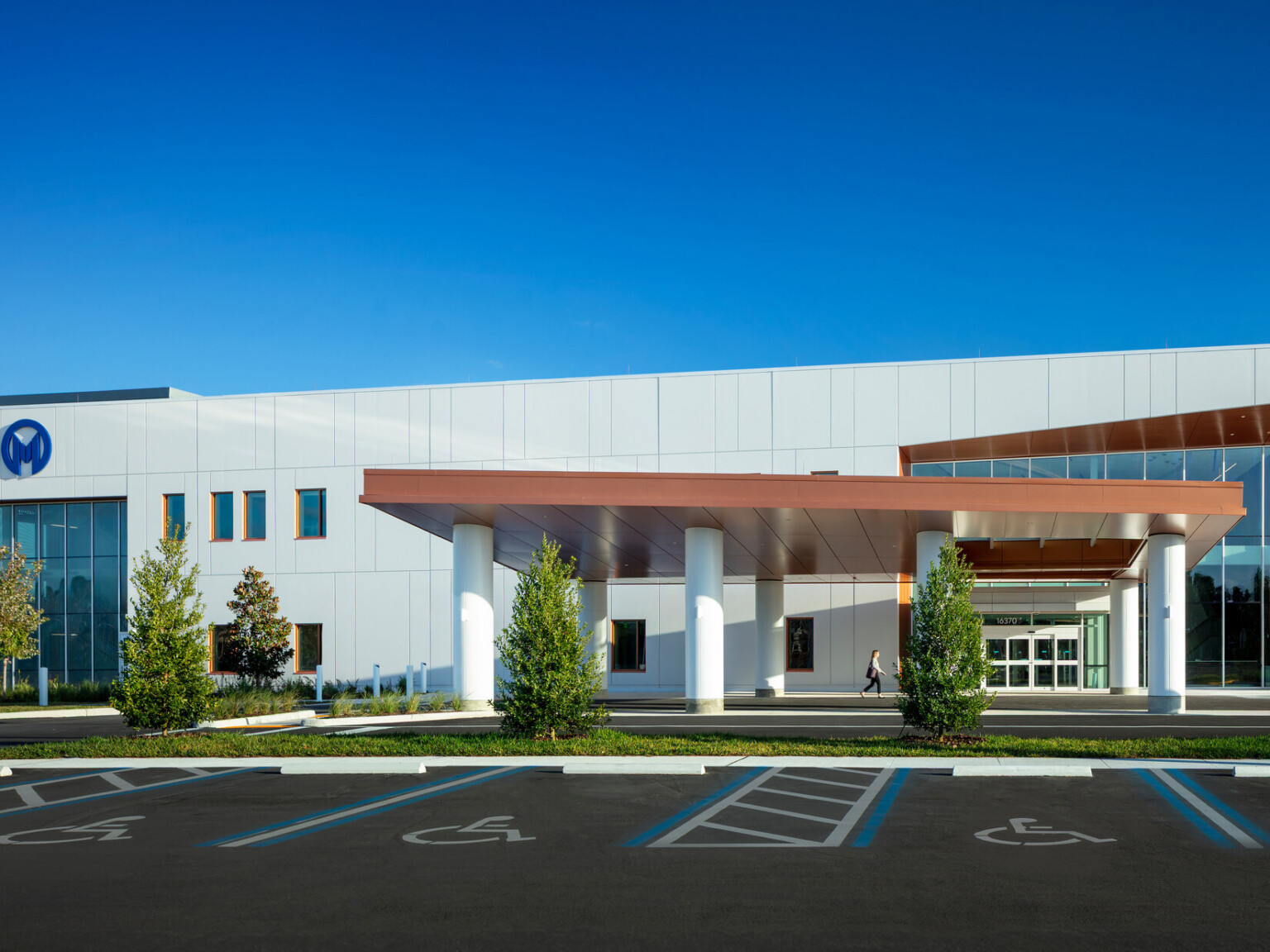 A modern building with a wide covered entrance, landscaped trees, and accessible parking spaces in the foreground under a bright blue sky