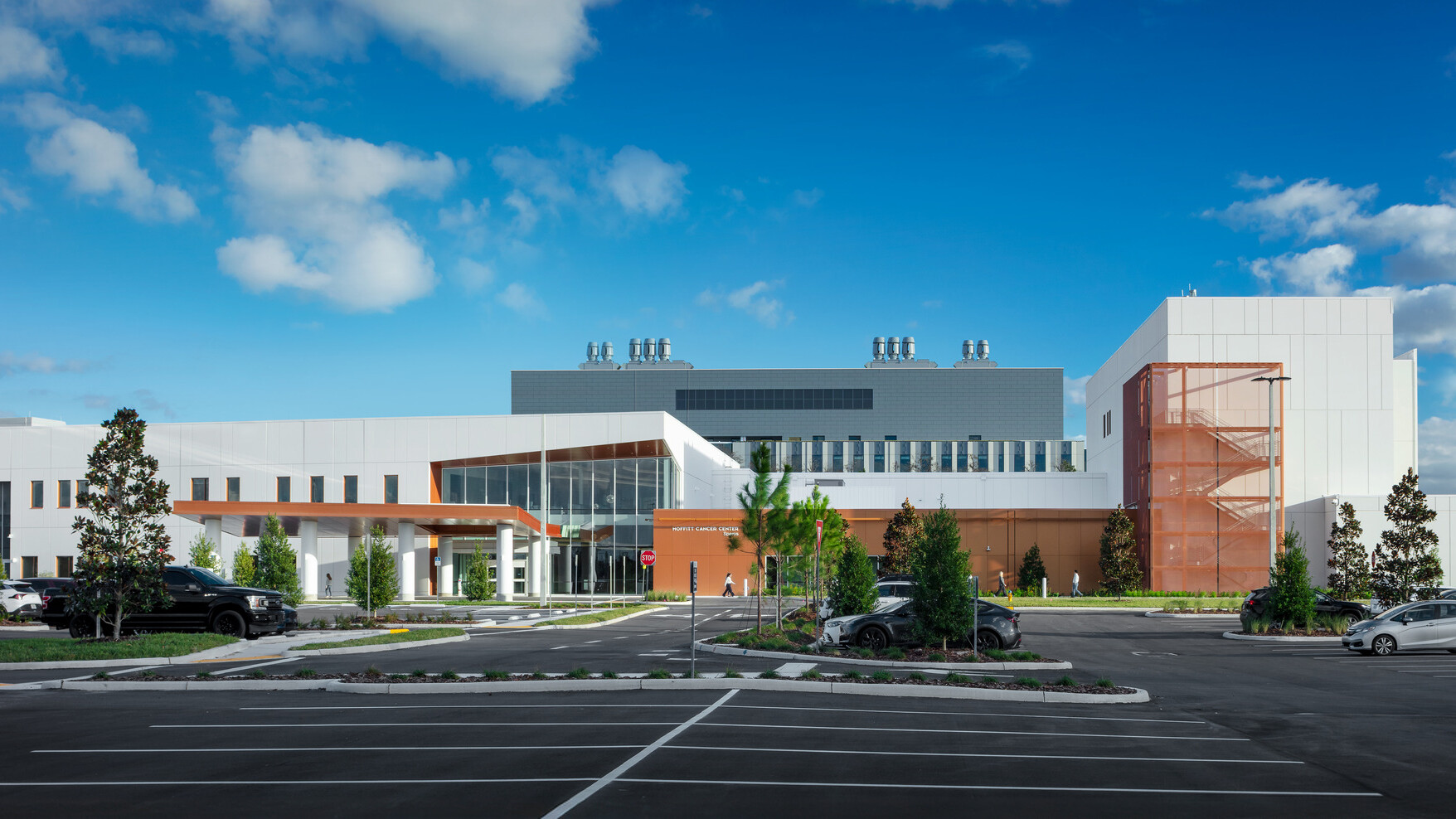 A modern, two-story building with a white façade, brown accents, and a large blue 'M' logo on the upper left corner. The building has several windows and a covered entrance area with two white vehicles parked nearby. Green landscaping and trees surround the building under a clear blue sky.