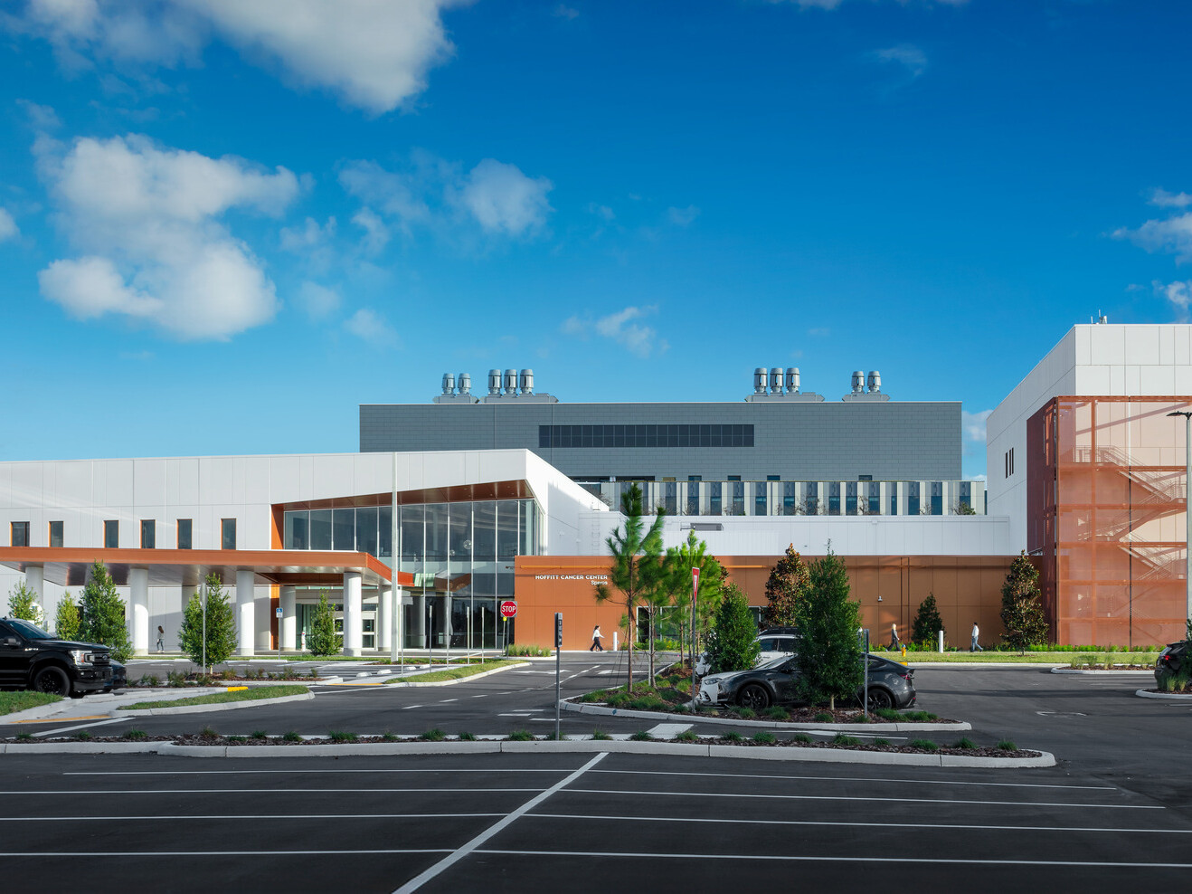 A modern, two-story building with a white façade, brown accents, and a large blue 'M' logo on the upper left corner. The building has several windows and a covered entrance area with two white vehicles parked nearby. Green landscaping and trees surround the building under a clear blue sky.