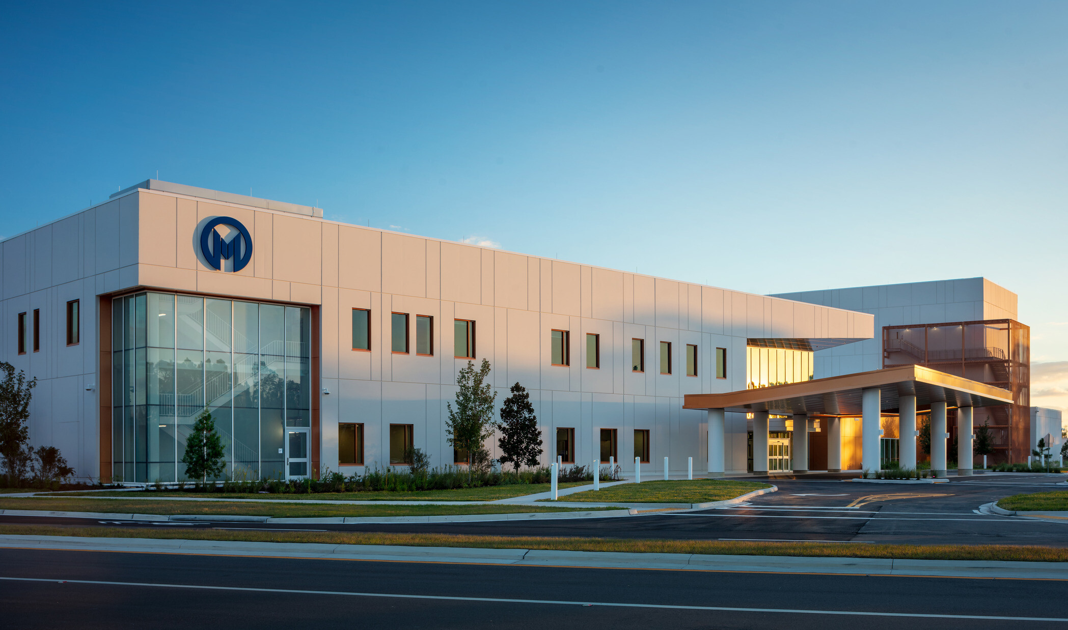 A modern, two-story building with a white façade, brown accents, and a large blue 'M' logo on the upper left corner. The building has several windows and a covered entrance area with two white vehicles parked nearby. Green landscaping and trees surround the building under a clear blue sky.