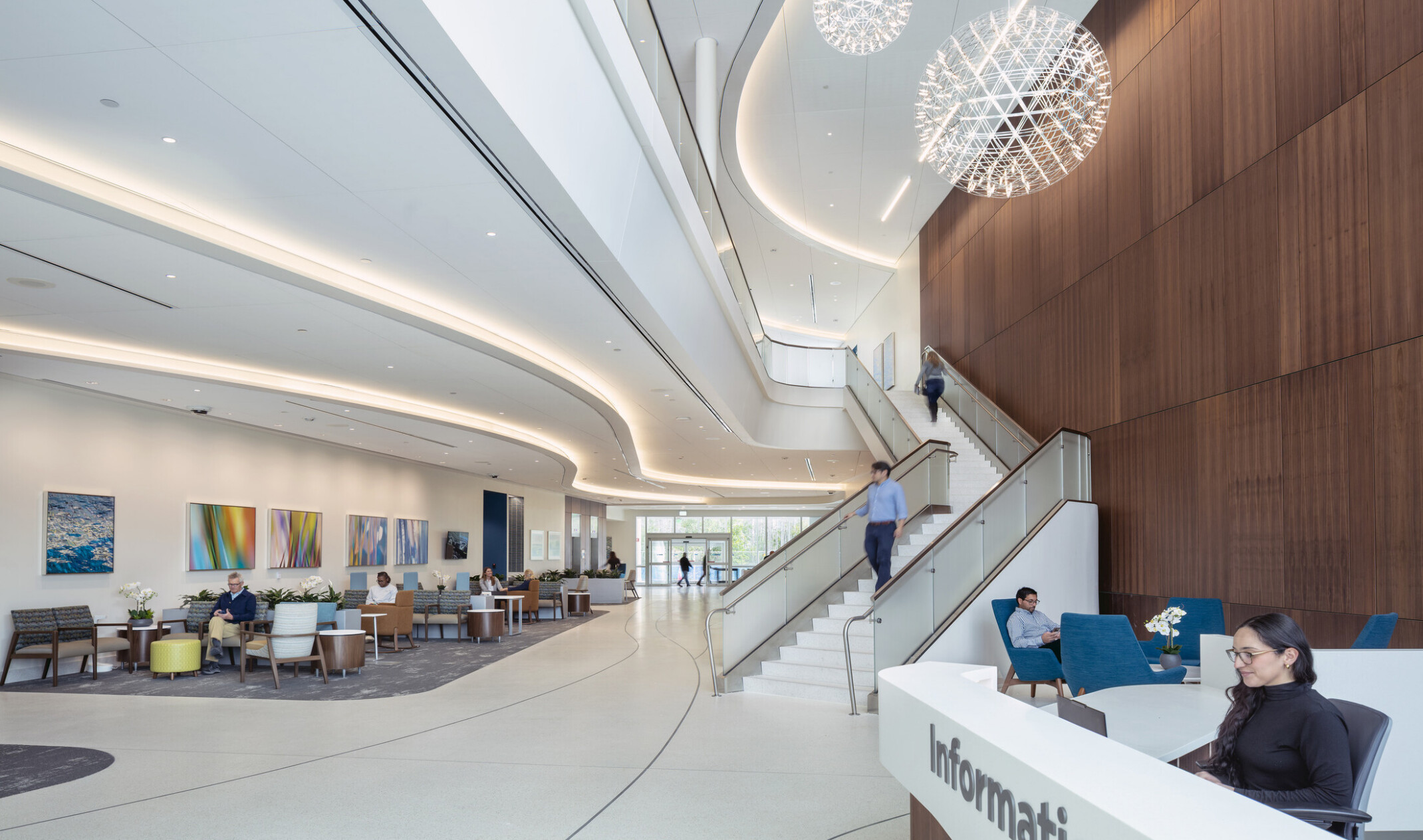 A modern, spacious lobby of the featuring a large central staircase, multiple reception desks with staff assisting visitors, and several people walking or sitting. The space includes wood-paneled walls, a vertical garden on the right, and polished terrazzo tile flooring.