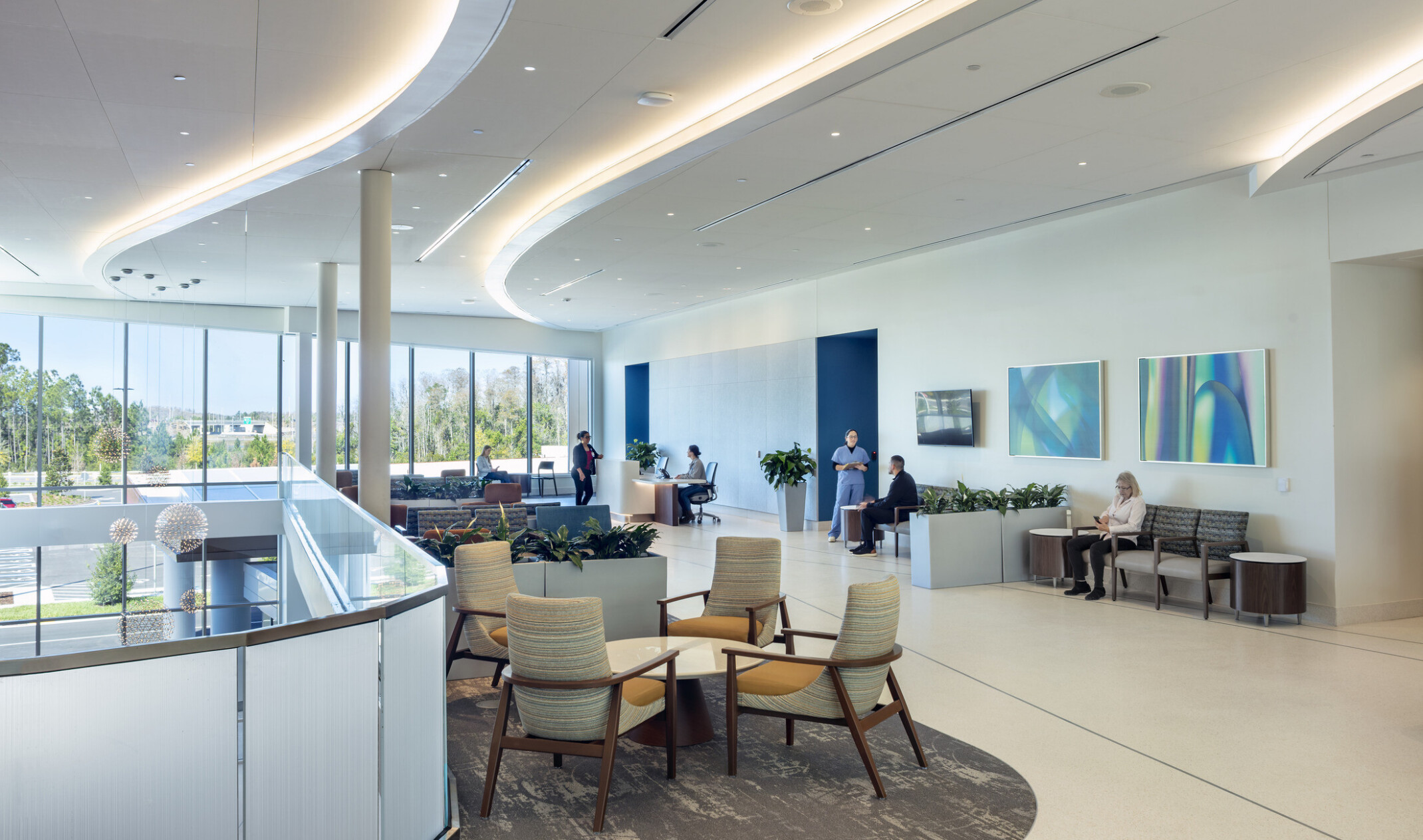 A modern, spacious lobby with a high ceiling and large windows that let in abundant natural light. The floor is made of polished stone tiles, and a glass railing lines a staircase leading to a lower level. People are walking, sitting in chairs, and standing near a reception desk.