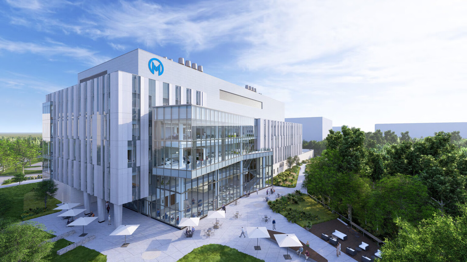 A modern, multi-story medical building with a glass façade and a logo featuring the letter 'M' on the top left corner. The building is surrounded by greenery and an outdoor courtyard with white umbrella-covered seating areas. People are walking and sitting in the courtyard.