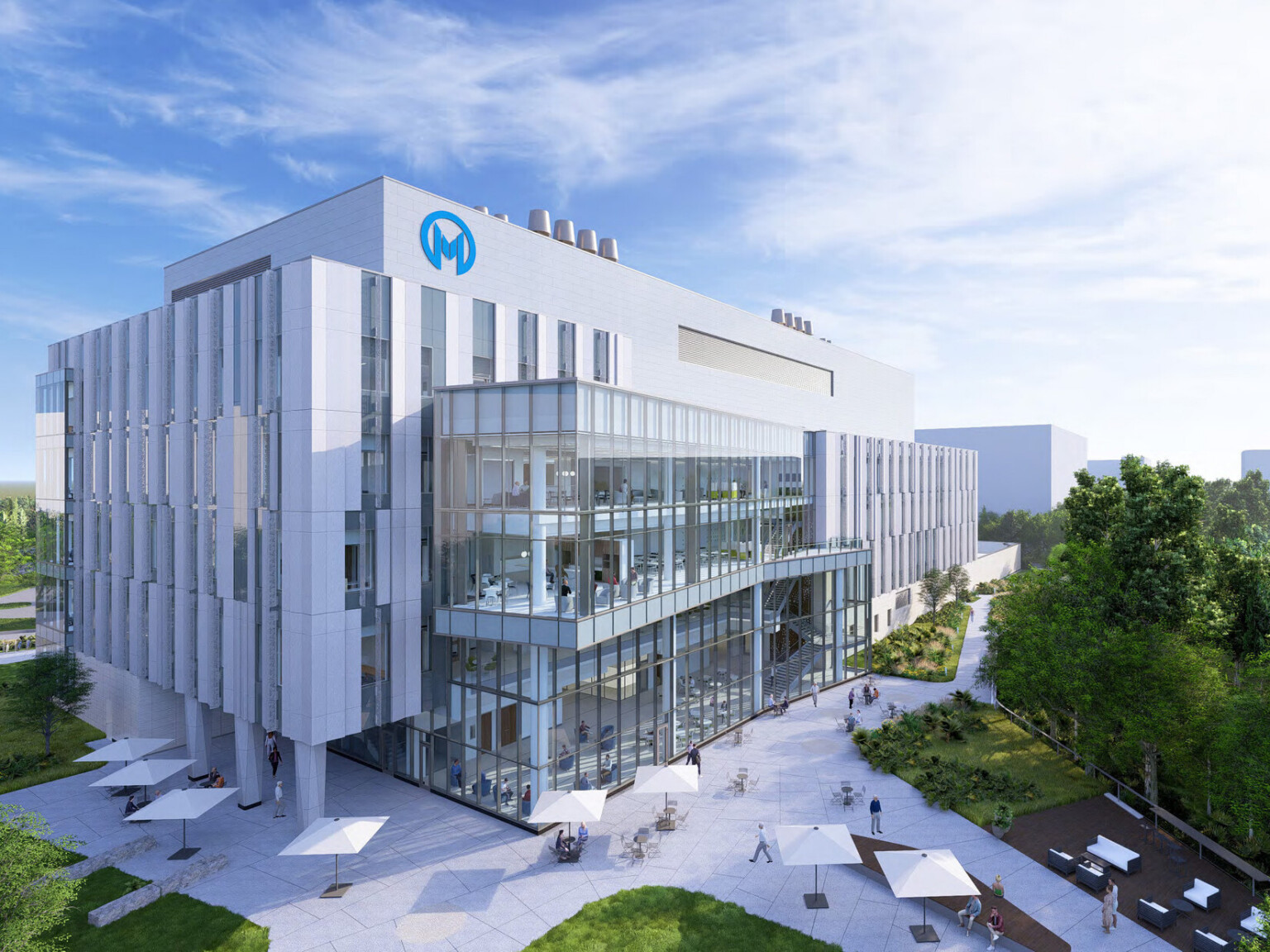 A modern, multi-story medical building with a glass façade and a logo featuring the letter 'M' on the top left corner. The building is surrounded by greenery and an outdoor courtyard with white umbrella-covered seating areas. People are walking and sitting in the courtyard.