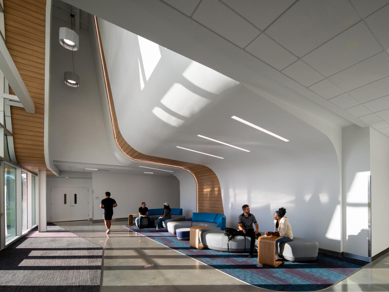 Modern interior space with high ceilings, large windows, and people sitting on blue and gray furniture. The white walls feature wooden accents curving along the ceiling.