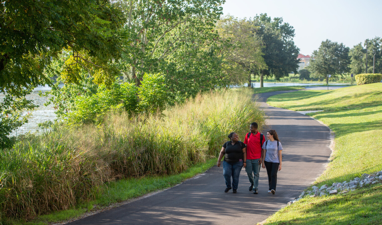 Three people walking on a curving paved path through a park with trees and grassy areas.