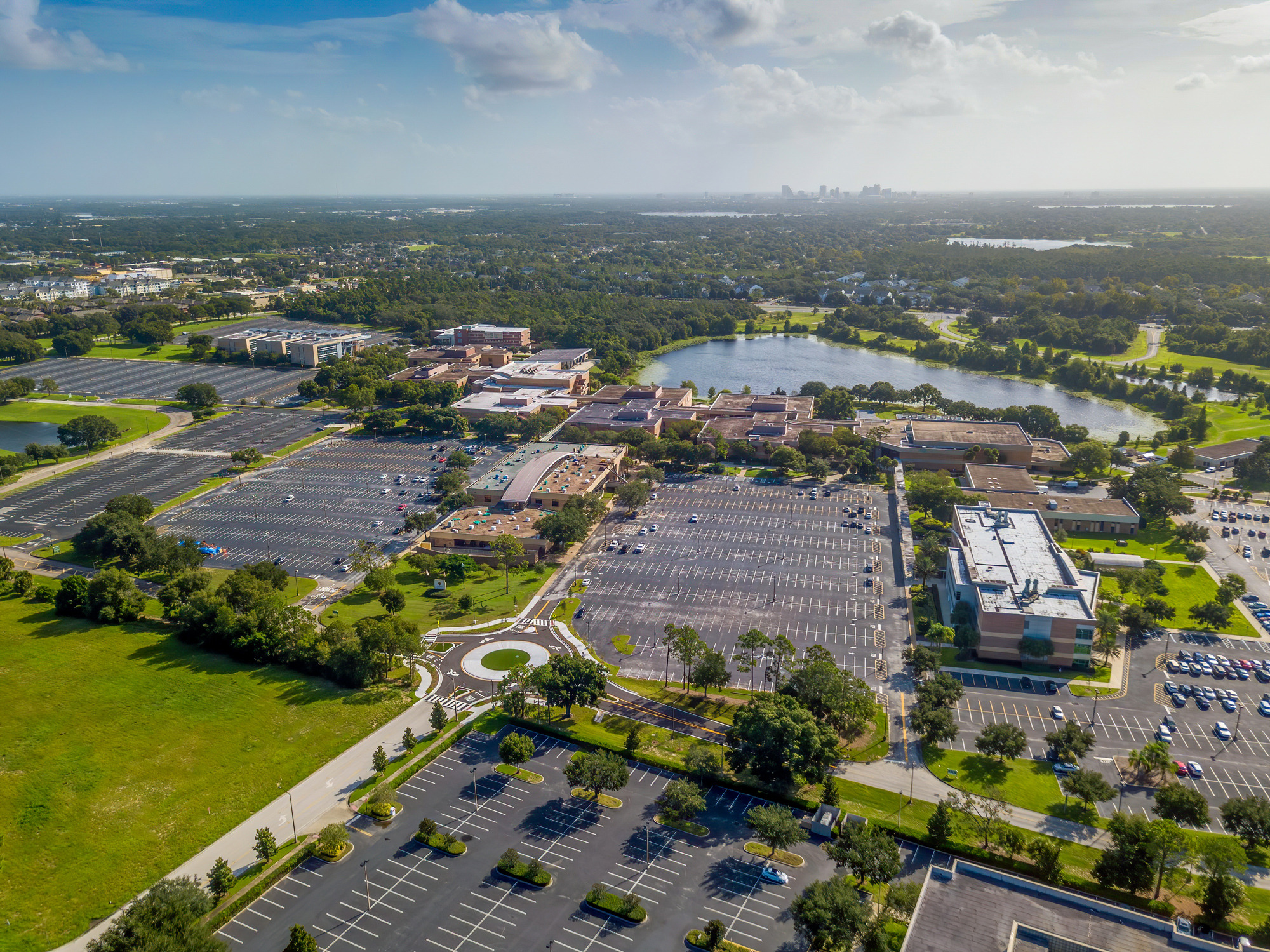 Aerial view of a campus with large buildings, parking lots, greenery, and small lakes, with a city skyline on the horizon.