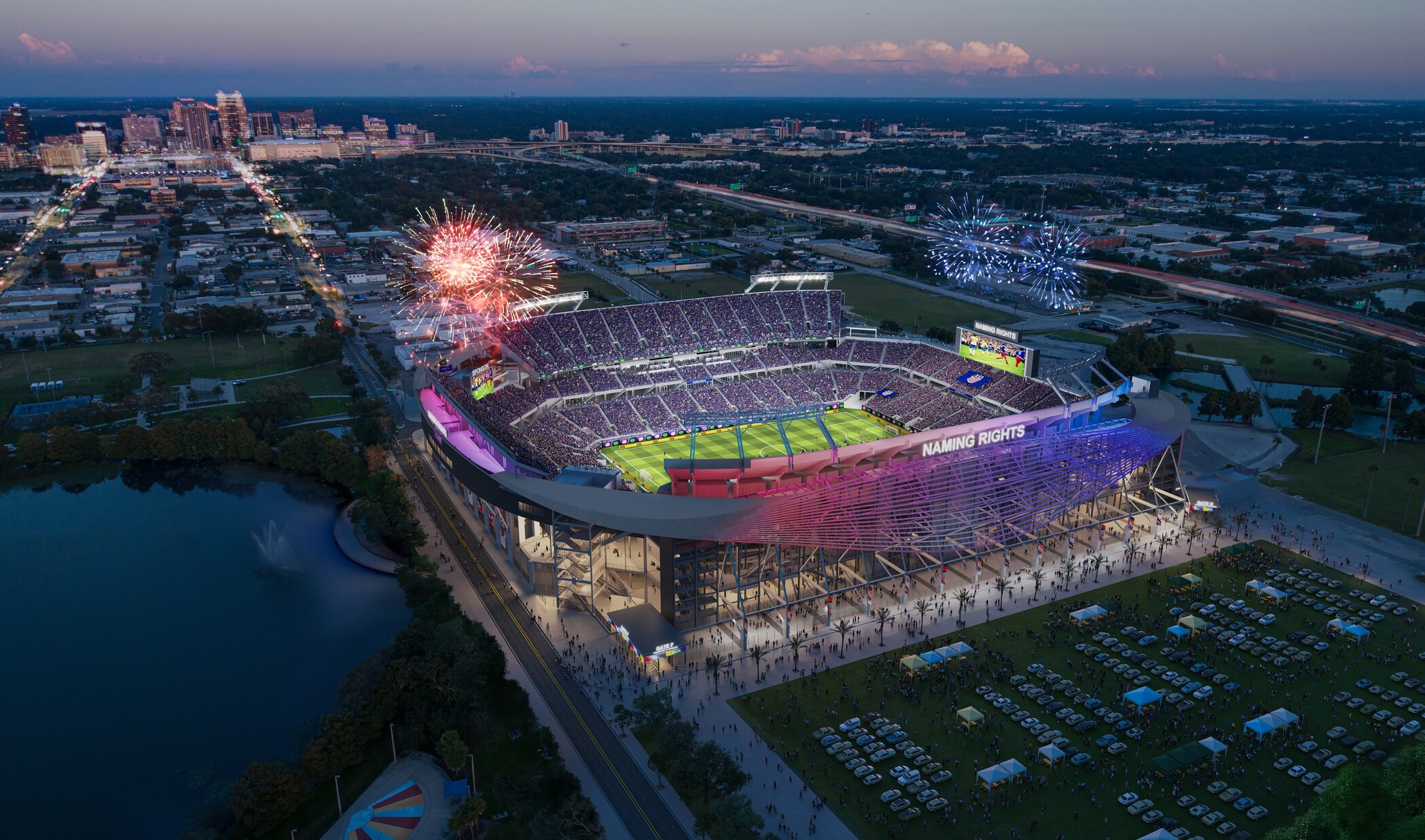 Aerial view of a large, brightly lit outdoor stadium at dusk, surrounded by parking lots, roads, and nearby buildings. Fireworks are exploding in multiple colors above