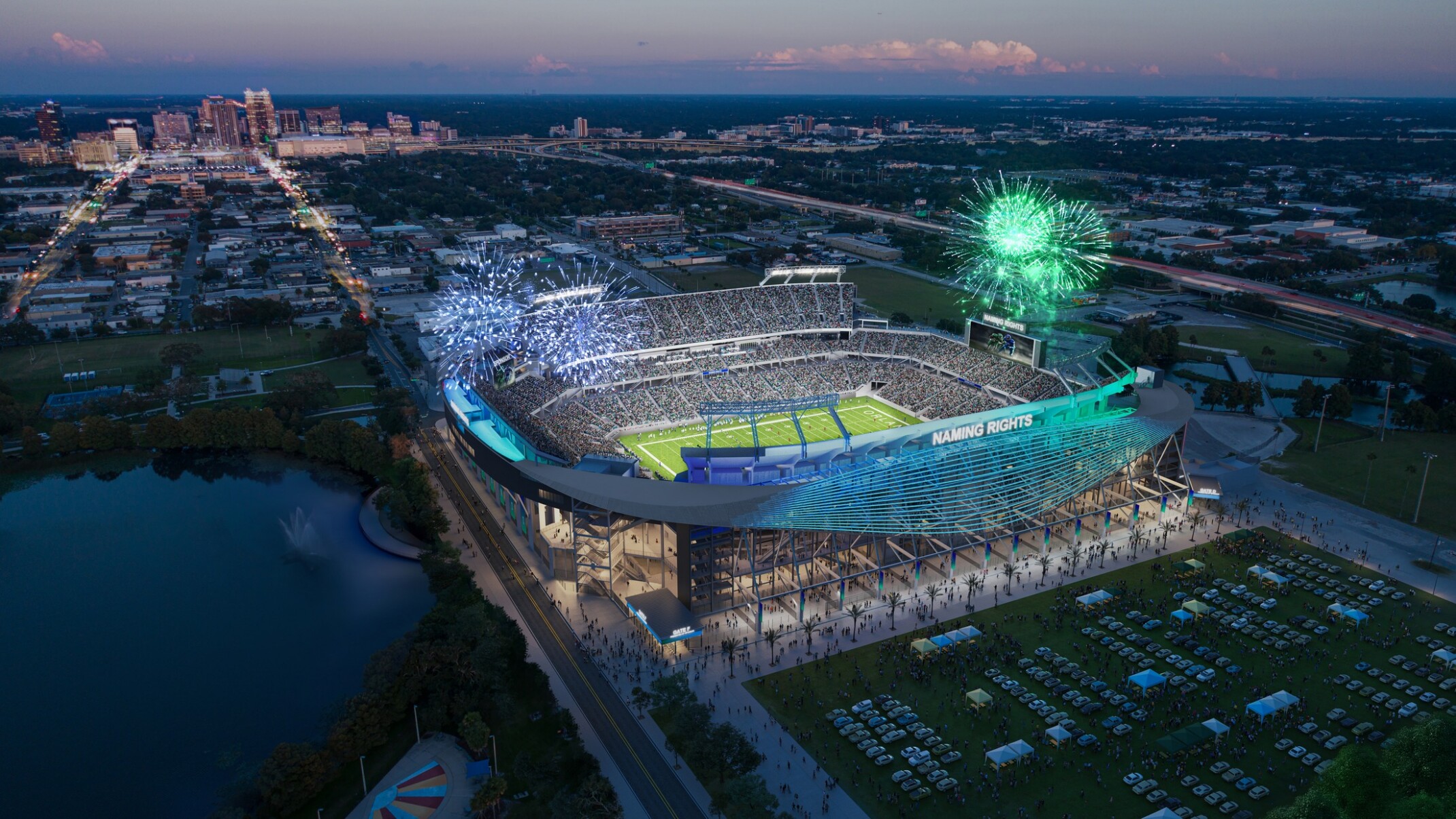 Aerial view of a large, brightly lit outdoor stadium at dusk, surrounded by parking lots, roads, and nearby buildings. Fireworks are exploding in multiple colors above