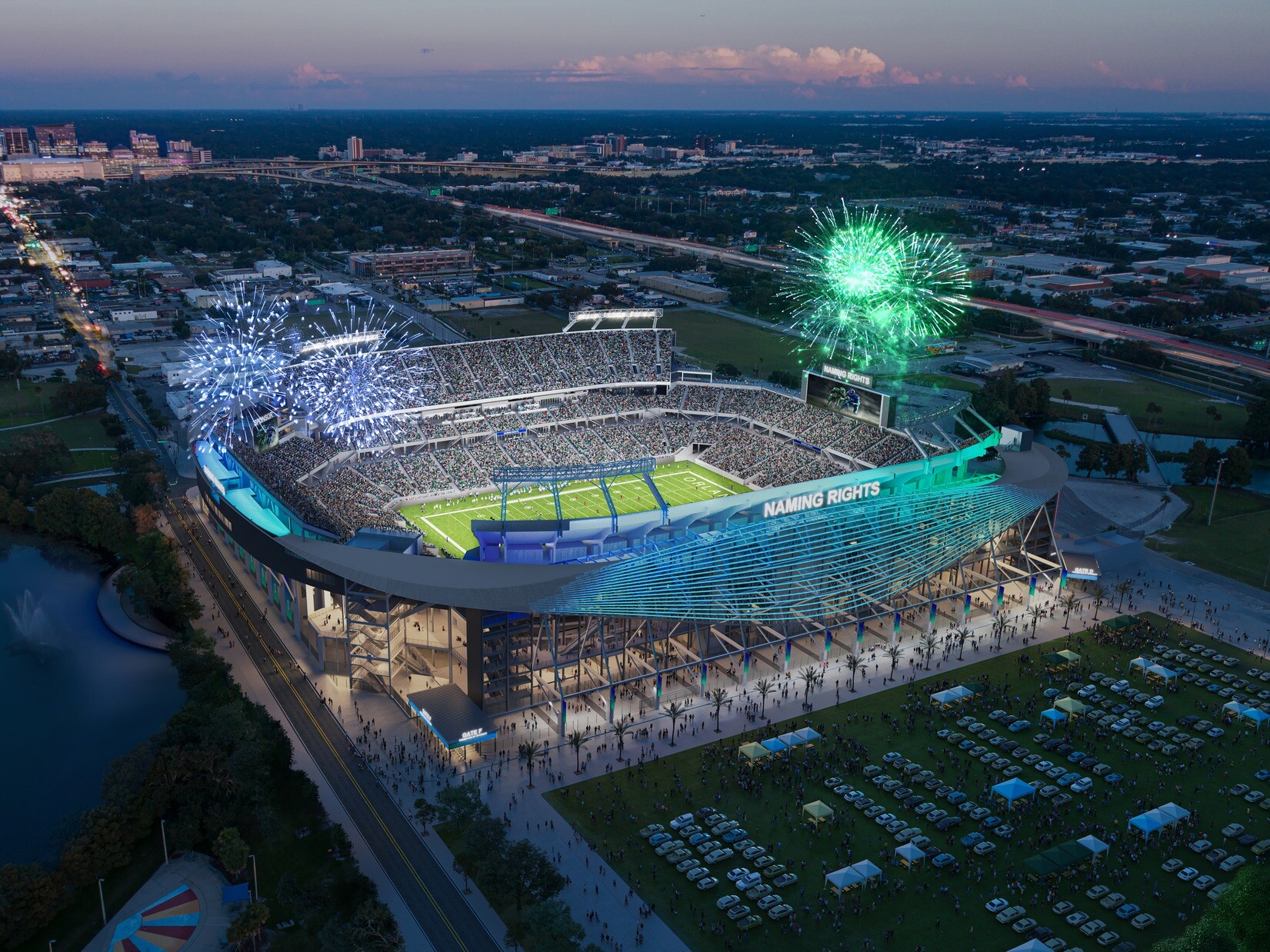 Aerial view of a large, brightly lit outdoor stadium at dusk, surrounded by parking lots, roads, and nearby buildings. Fireworks are exploding in multiple colors above