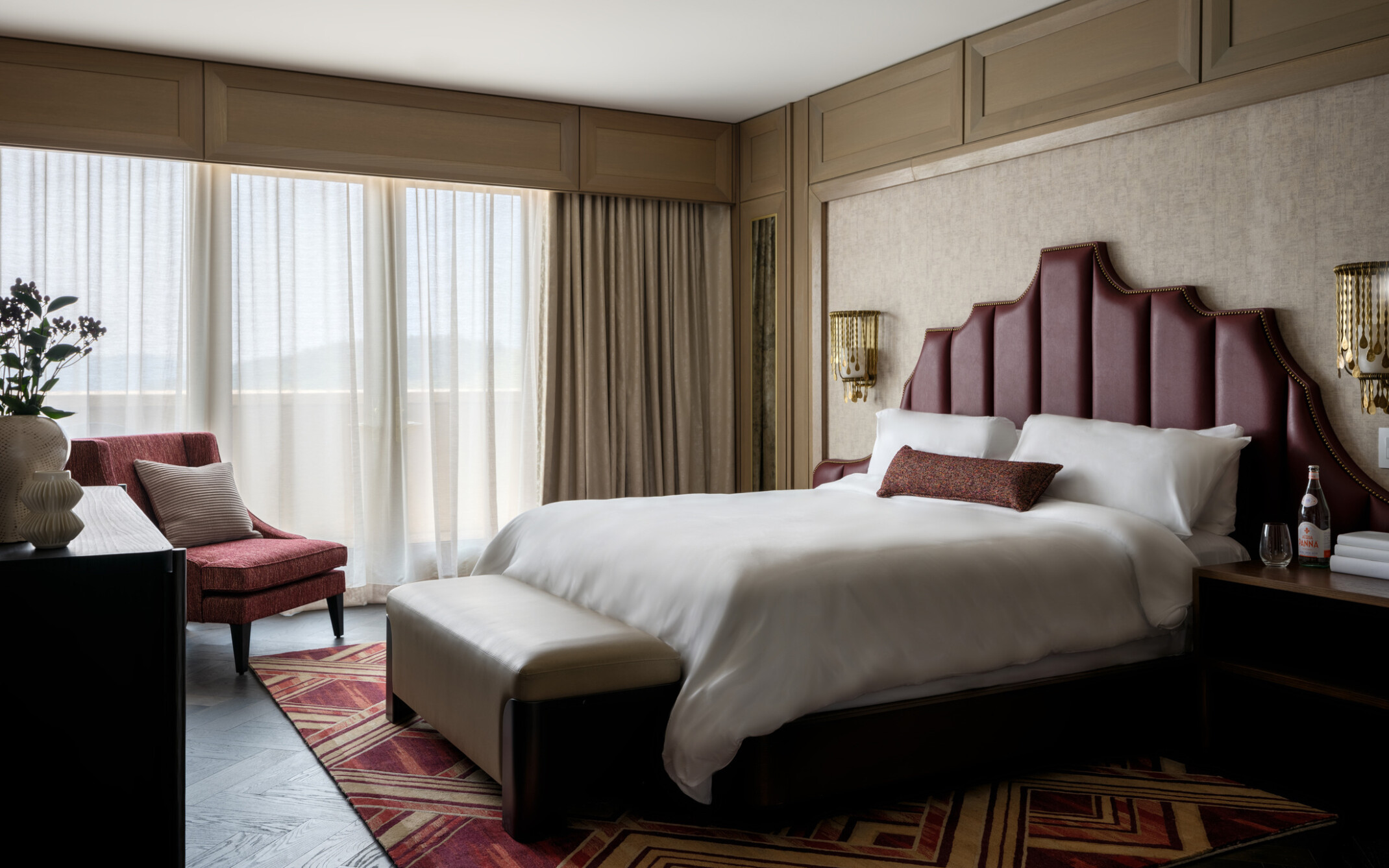 Hotel room with bed with red headboard and soft white linens, red accent chair, large windows covered by sheer curtains