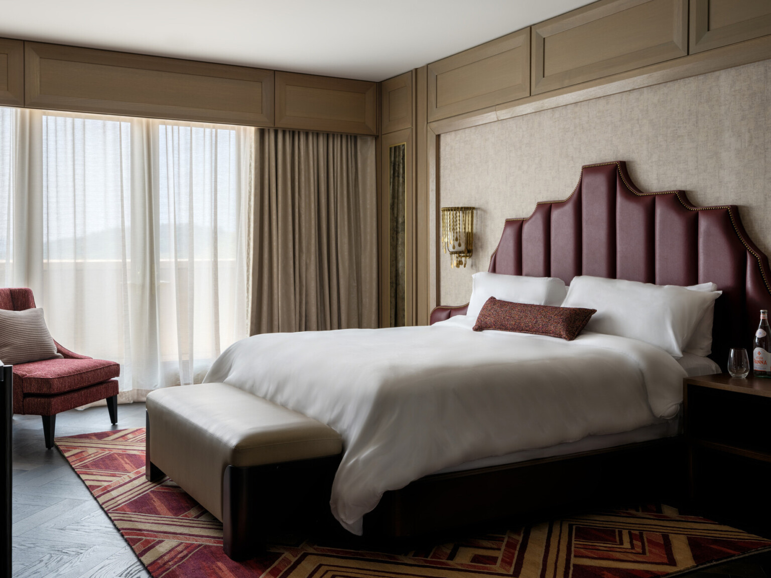 Hotel room with bed with red headboard and soft white linens, red accent chair, large windows covered by sheer curtains