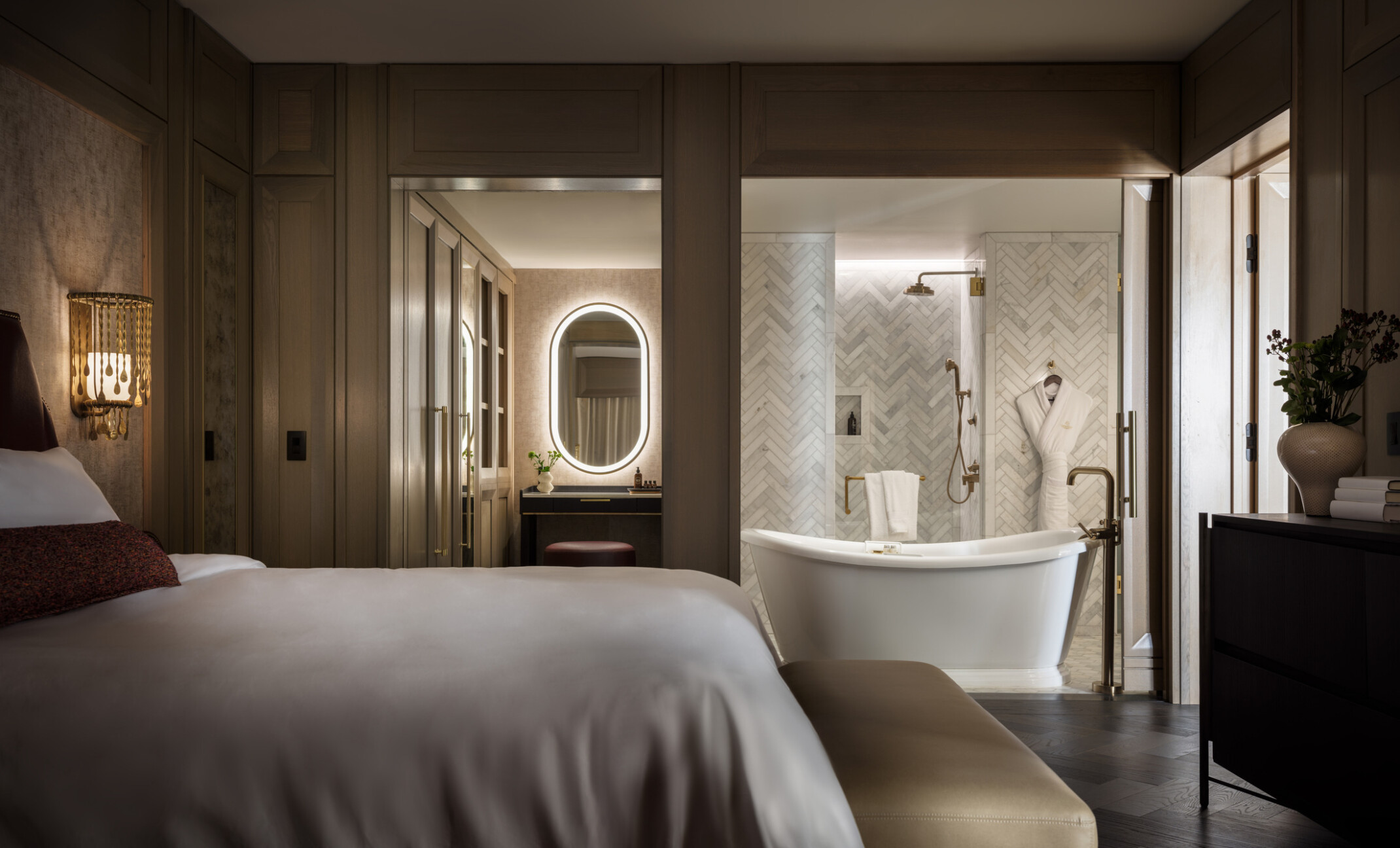Dimly lit hotel room showcasing a bathroom with large tub in front of a grey and white tile wall, oval lit mirror, bed with soft white linens Dimly lit hotel room showcasing a bathroom with large tub in front of a grey and white tile wall, oval lit mirror, bed with soft white linens