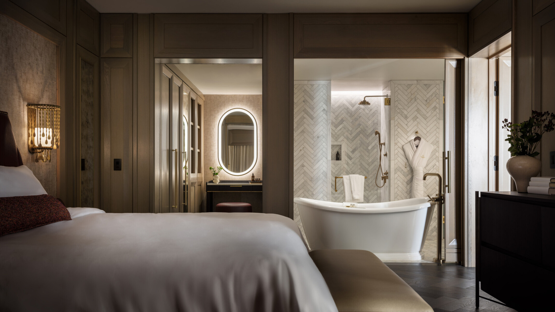 Dimly lit hotel room showcasing a bathroom with large tub in front of a grey and white tile wall, oval lit mirror, bed with soft white linens