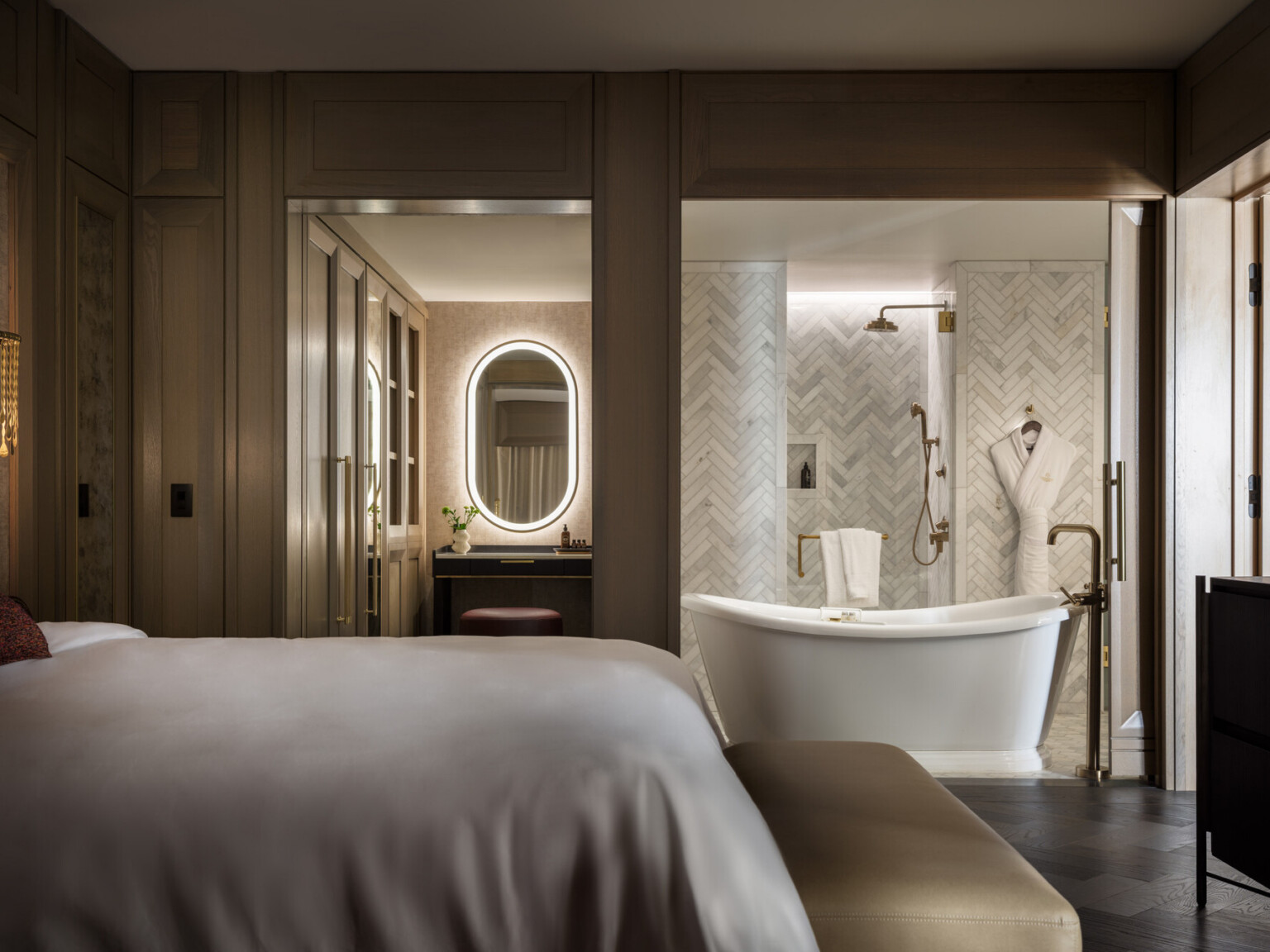Dimly lit hotel room showcasing a bathroom with large tub in front of a grey and white tile wall, oval lit mirror, bed with soft white linens