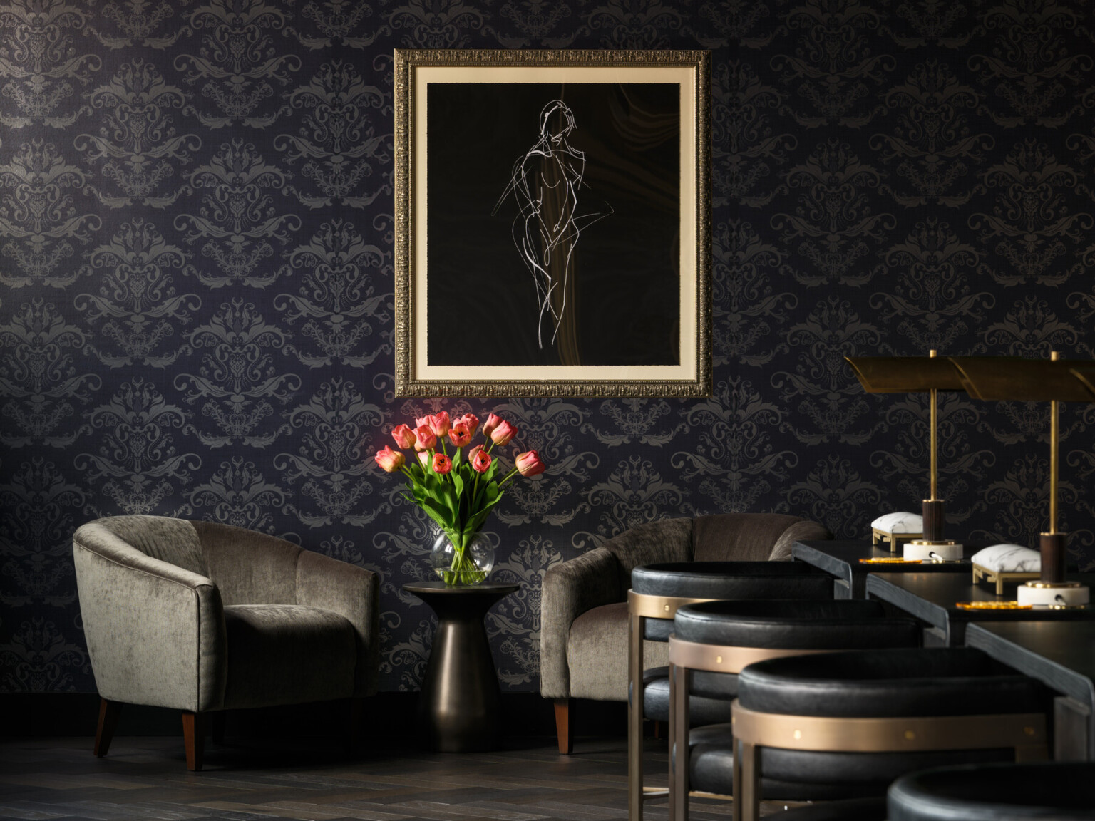 Bar seating area with gold and black bar stools, black and grey wall paper, grey velvet chairs with artwork hanging on the wall above