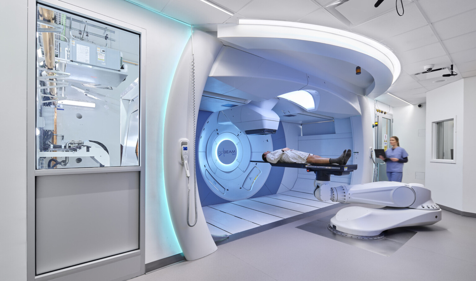 Modern medical facility with a patient on a robotic treatment table in front of a large circular machine, assisted by a medical professional.