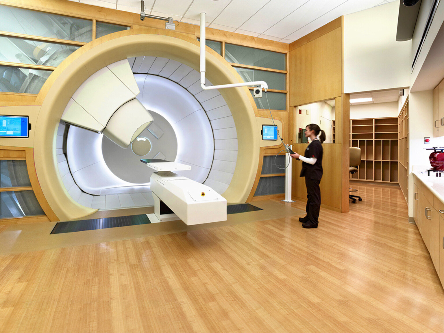 Medical room with a large circular imaging or radiation machine, monitors, and a staff member interacting with equipment.