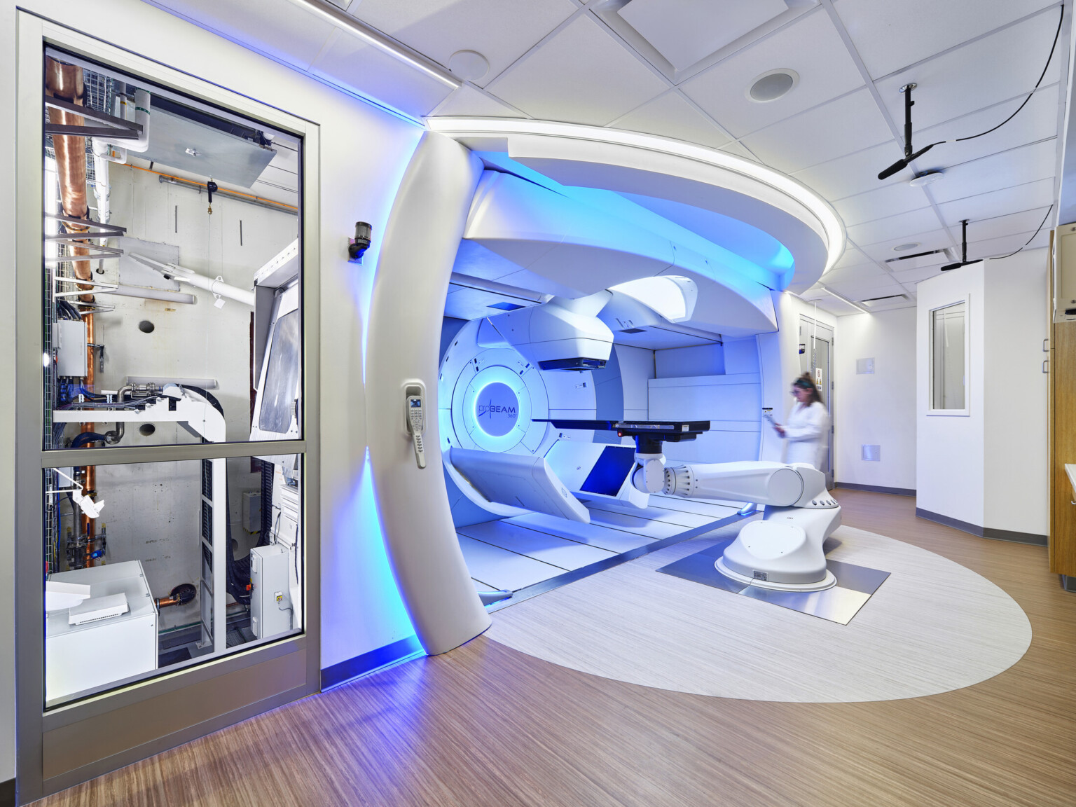 Medical treatment room featuring a modern proton therapy machine, illuminated with blue accent lighting. A healthcare professional in a white lab coat is seen blurred in motion near the machine.