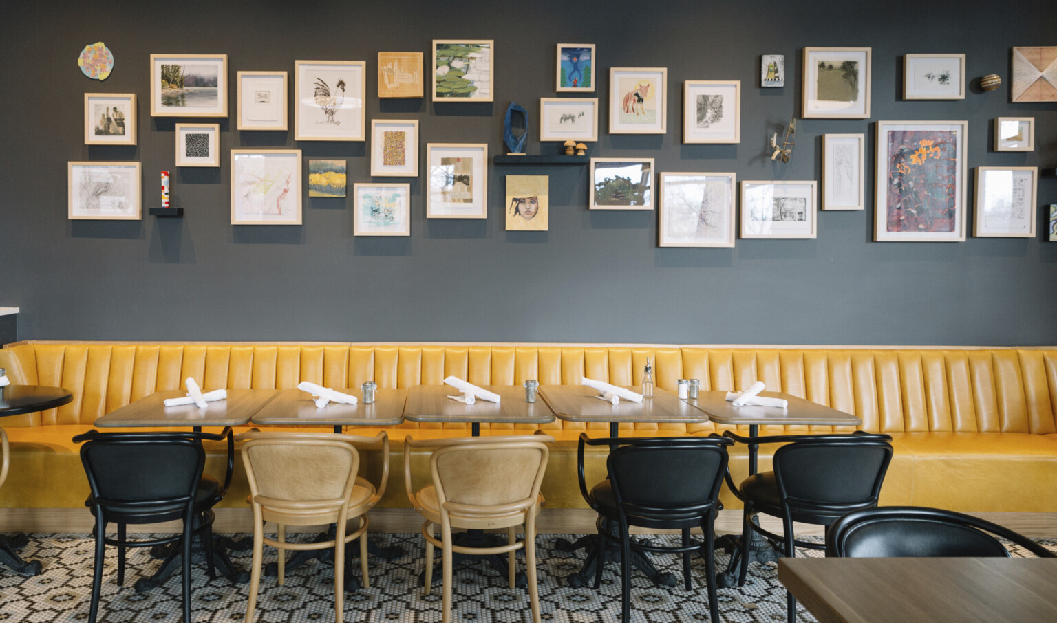 Yellow booth lines dark blue grey wall with gallery style art collage. Wood tables with black and brown chairs in front