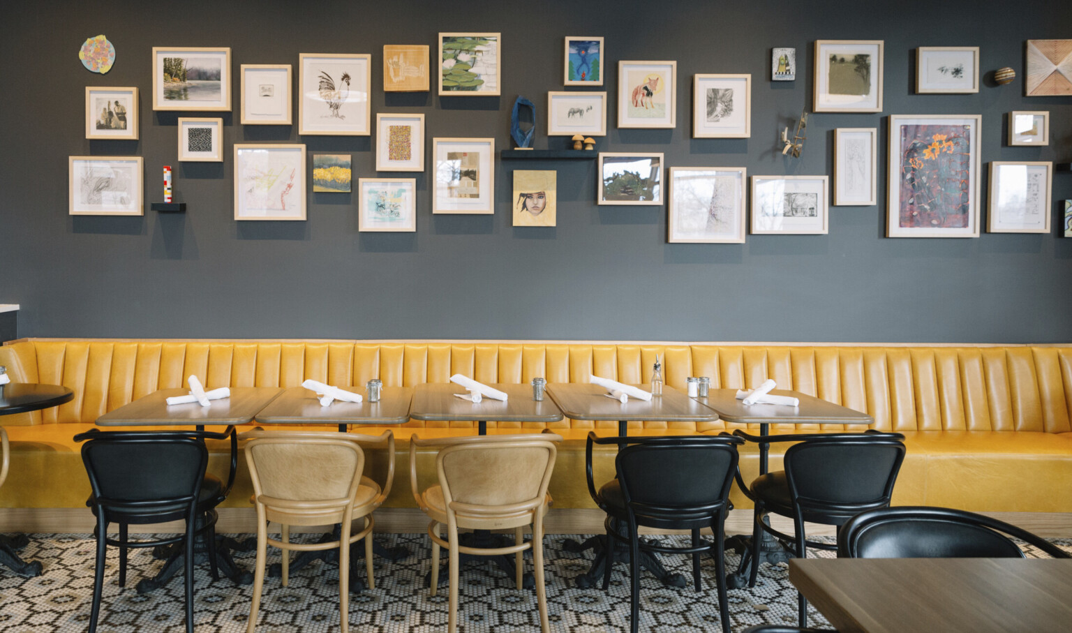 Yellow booth lines dark blue grey wall with gallery style art collage. Wood tables with black and brown chairs in front