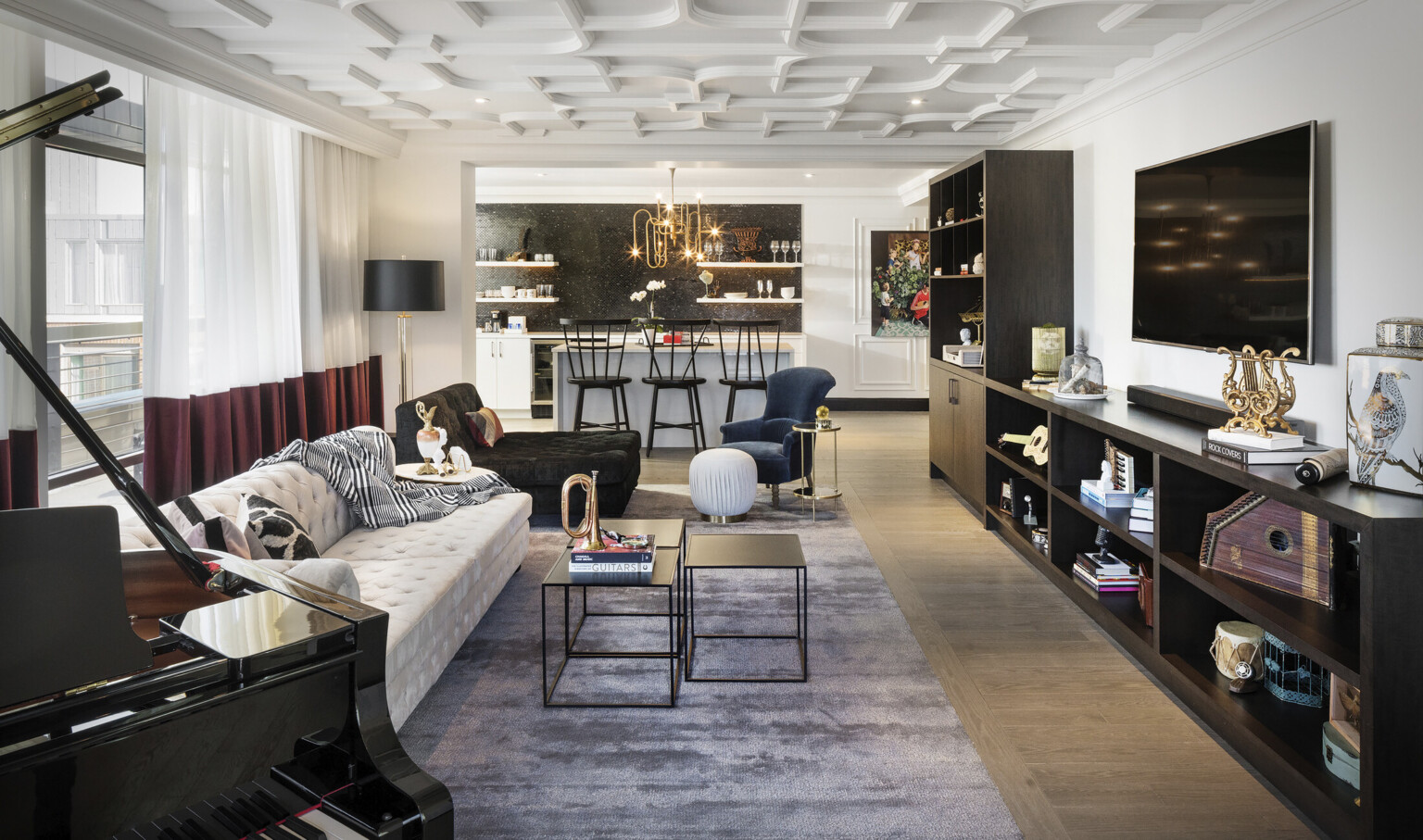 White living room with seating area facing dark wood shelves under screen. Beyond is white kitchen with island and black tile