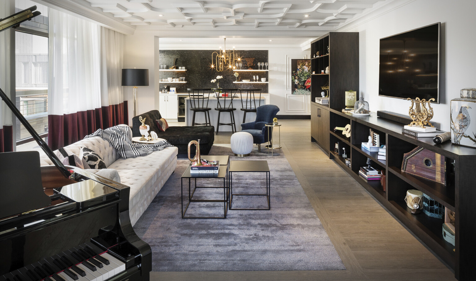 White living room with seating area facing dark wood shelves under screen. Beyond is white kitchen with island and black tile