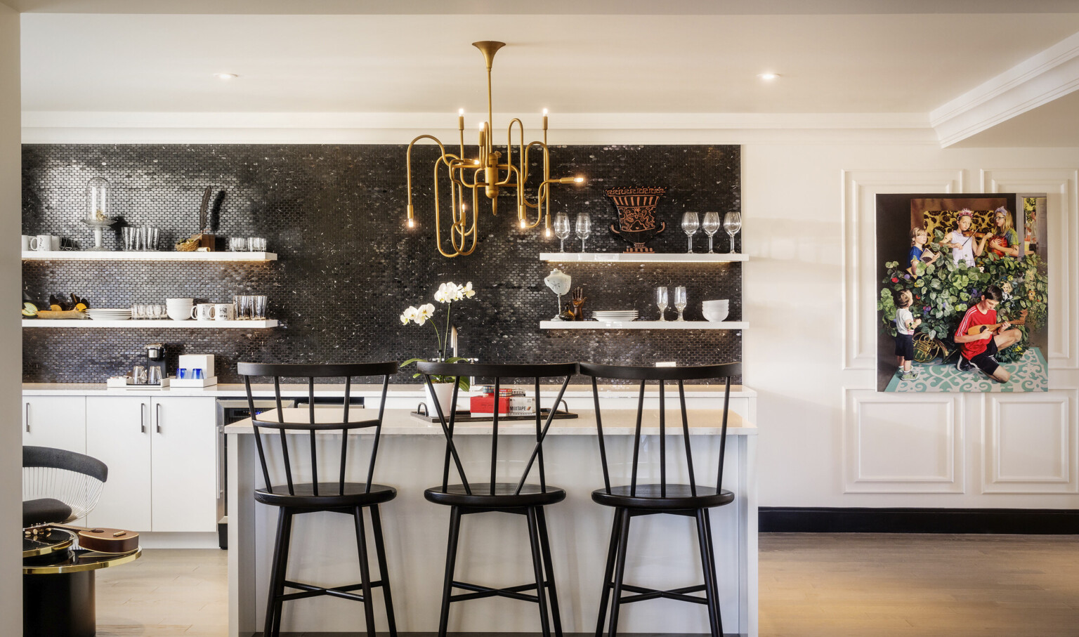 Closeup of kitchen. Black tile accent over white counter. A gold organically curved chandelier hangs over a white island