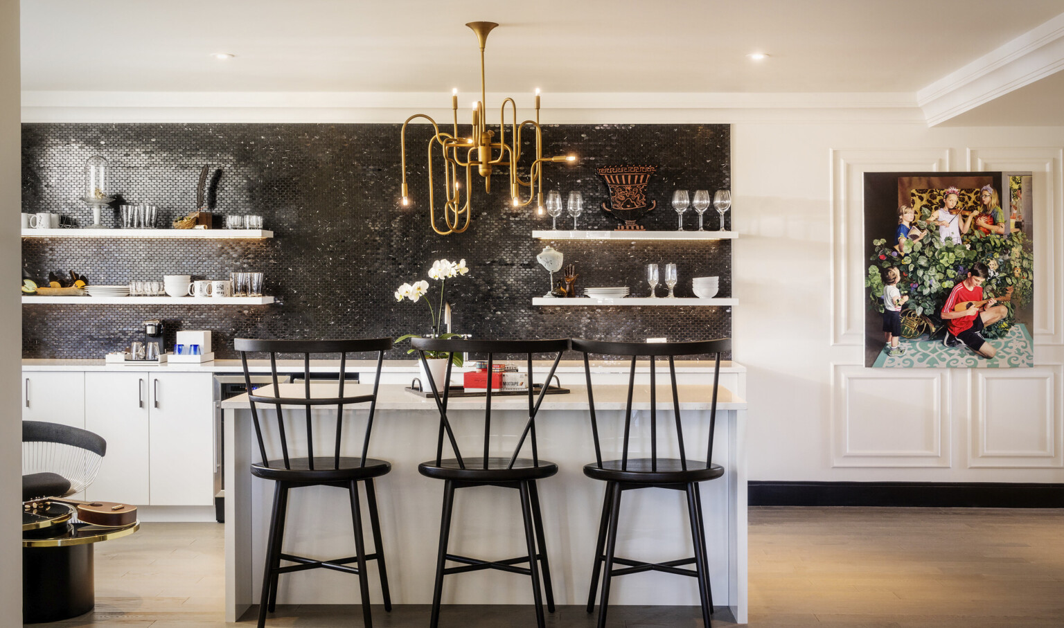 Closeup of kitchen. Black tile accent over white counter. A gold organically curved chandelier hangs over a white island