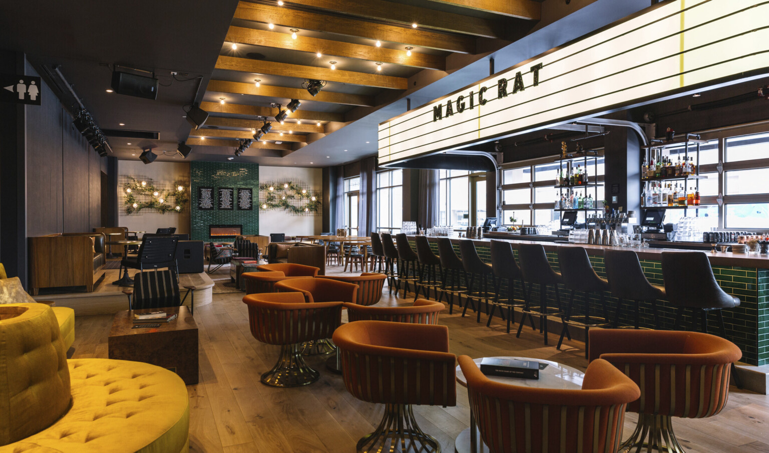 Green tiled bar with warm toned flexible seating area. Arm chairs and couches by green tiled fireplace framed by white walls