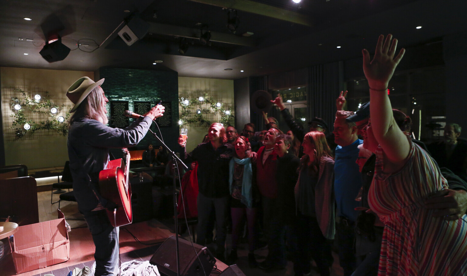Man performs for crowd in Magic Rat nightclub, green fireplace illuminated with greenery and lighting on white walls by fire