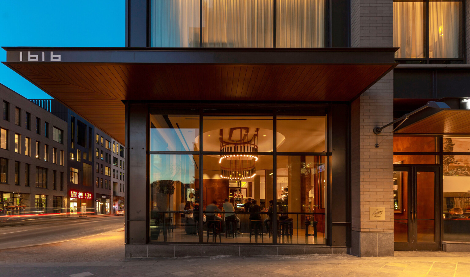 View through floor to ceiling window to street level lounge. Black canopy extends over sidewalk, wrapping around corner