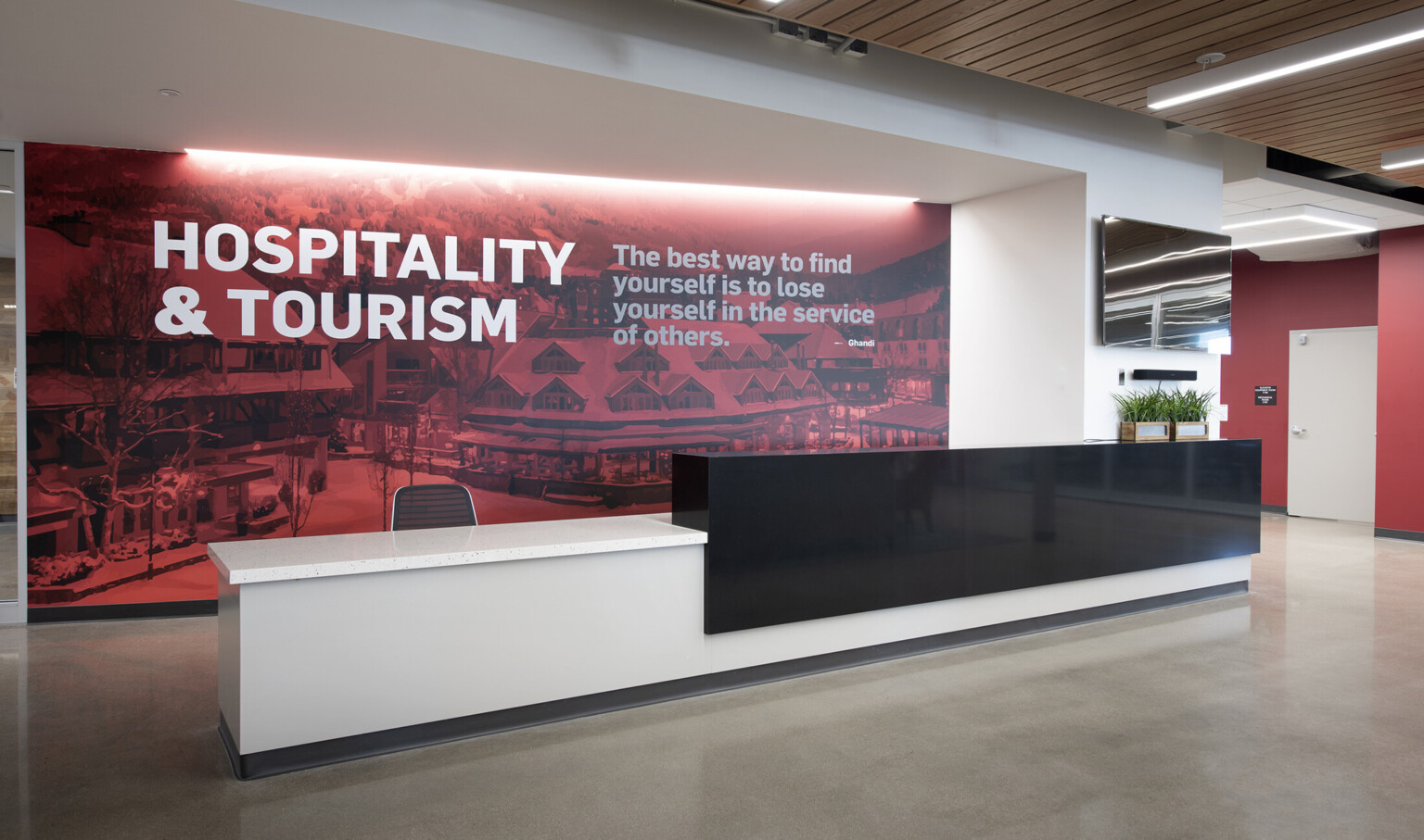White desk with black partition in front of recessed wall mural with writing: Hospitality and Tourism, and quote from Ghandi