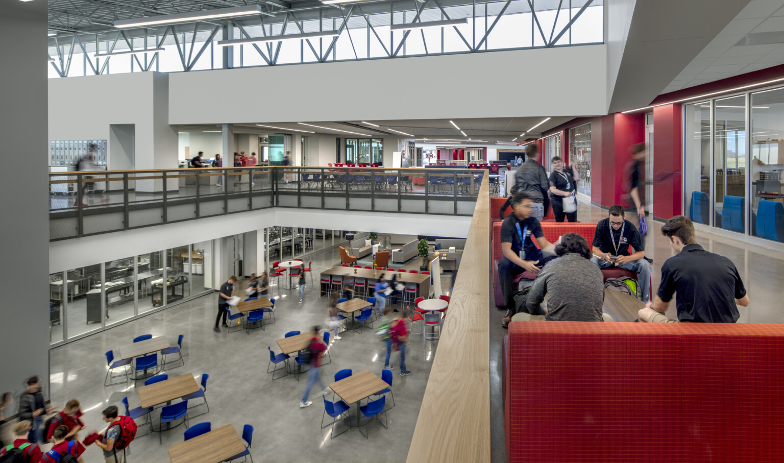 View of a common area filled with tables and blue chairs from the second floor standing by a red couch in a area filled with students