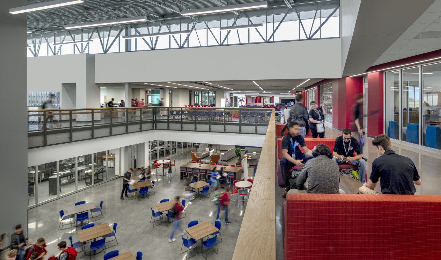 View of a common area filled with tables and blue chairs from the second floor standing by a red couch in a area filled with students