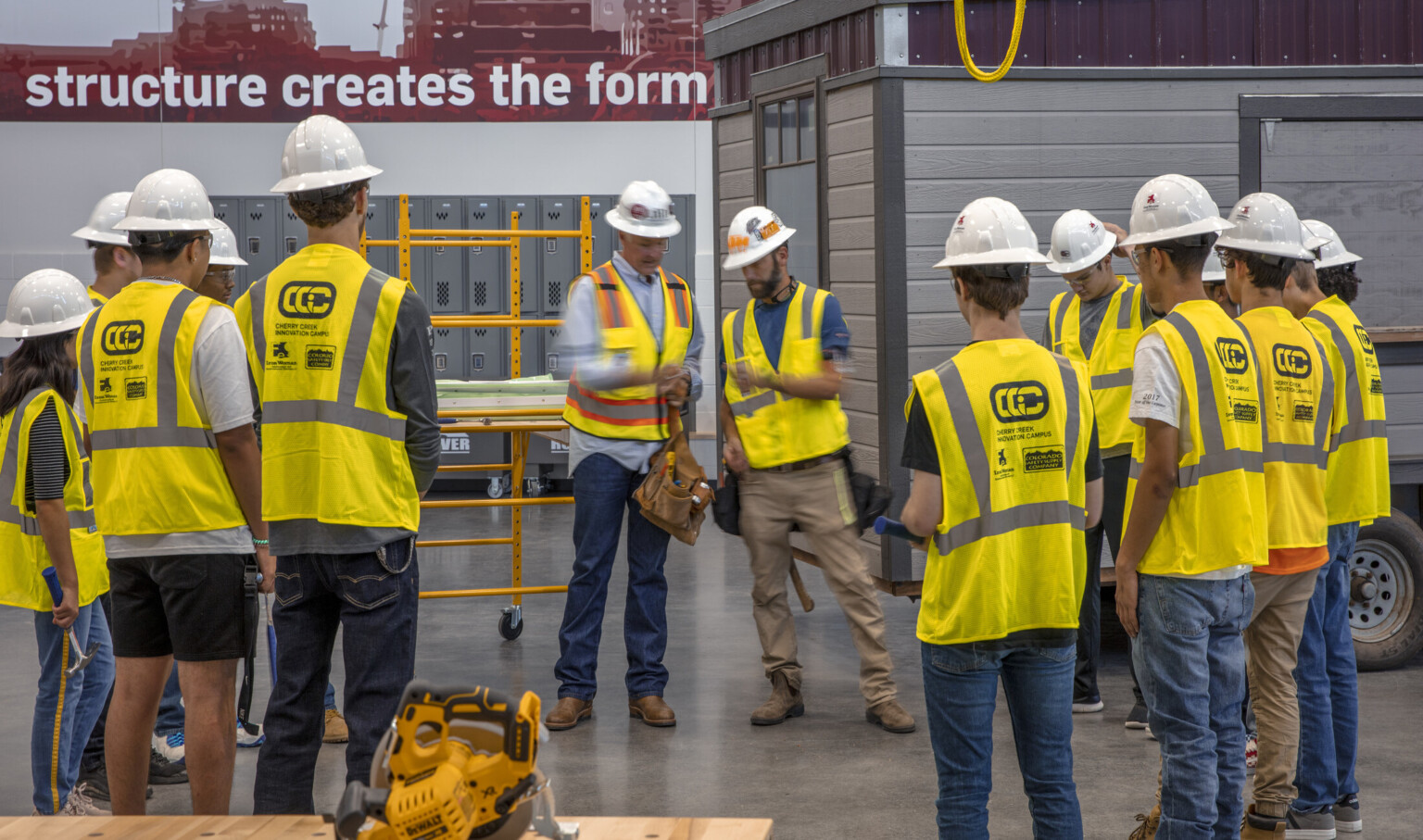 Circular saw on table in front of students in safety vest and hard hats circled around teachers. Paneled room, back right