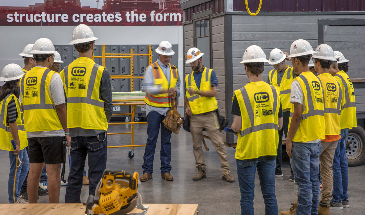 Circular saw on table in front of students in safety vest and hard hats circled around teachers. Paneled room, back right Circular saw on table in front of students in safety vest and hard hats circled around teachers. Paneled room, back right