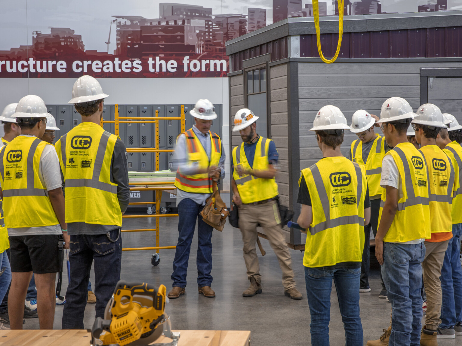 Circular saw on table in front of students in safety vest and hard hats circled around teachers. Paneled room, back right