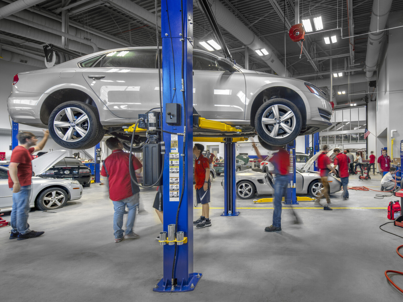 Students work on lifted car in double height mechanics lab. More cars on the ground behind. Exposed duct work on grey ceiling