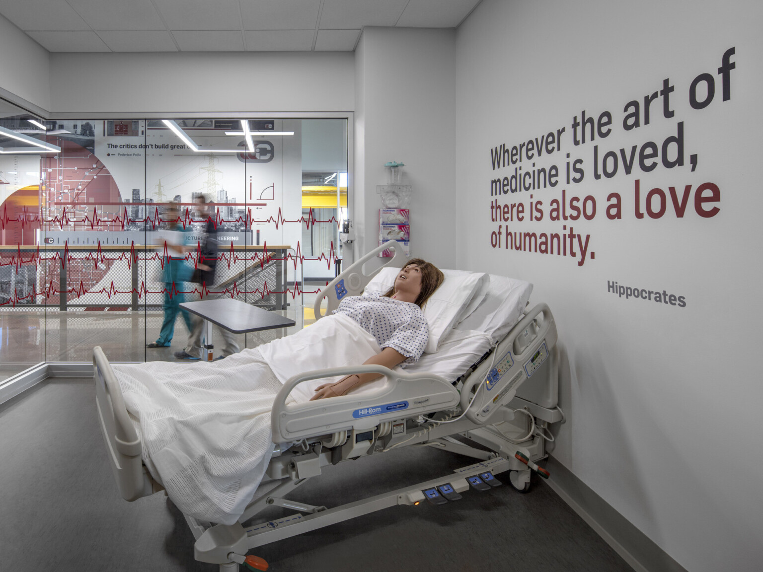 Health science lab with hospital bed and CPR dummy. Hippocrates quote on right wall. Glass walls to hall with zigzag design