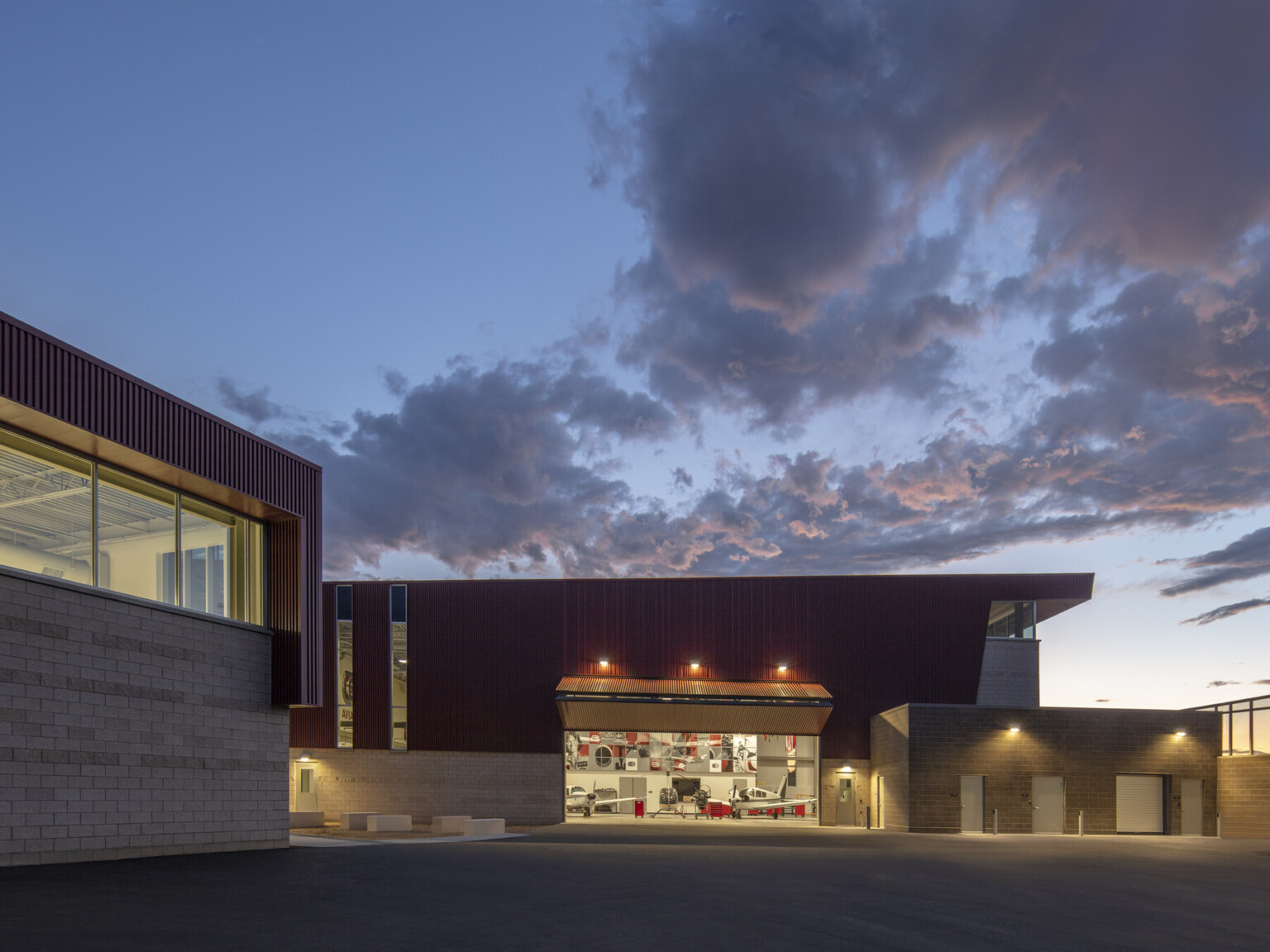 Stone building with textured red wrap facade illuminated from within in evening. Garage door opened at center to airplanes