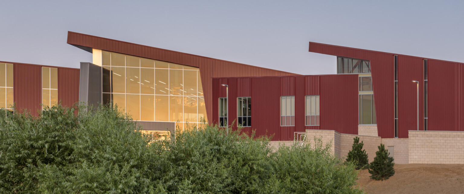 Stone base on building below textured red panel wrapped facade with angled roofs. Angled double height glass entrance