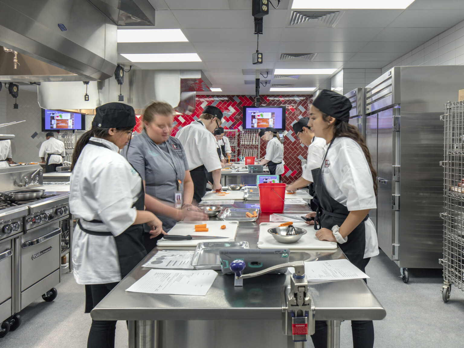 Culinary kitchen classroom with stainless steel island and red tile back wall. Stoves on left wall, refrigerators to right