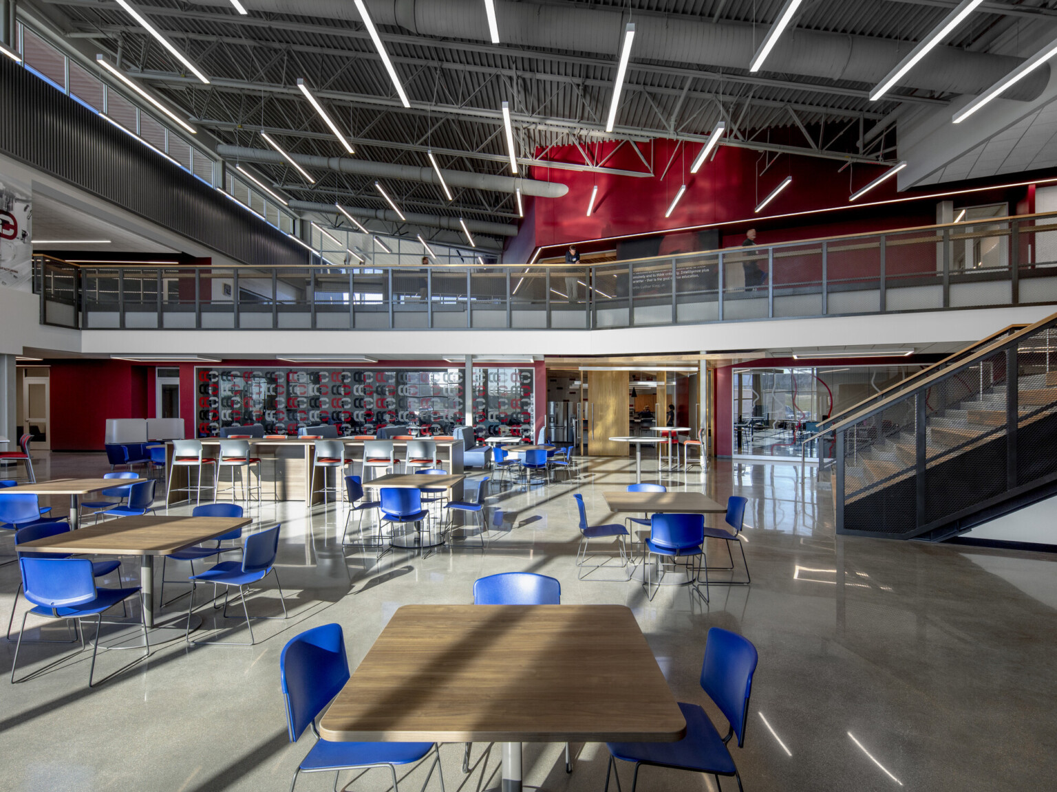 Seating area in double height room with natural lighting. 2nd floor walkway to back extends over entrance to classroom