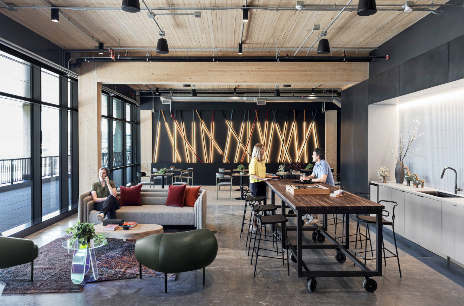 interior of a mass timber building; floor to ceiling windows with steel frame and mass timber elements exposed; gray concrete floor; people in a communal kitchen space with varied seating talking; Right wall with sink faces floor to ceiling windows with patio space; backwall lighting design elements feature long sticks of light laid on top of each other